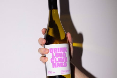 Hand holding wine bottle with bold pink label text Drink Loud Clink Hard perfect for graphic mockup design projects and branding inspiration дизайнеры