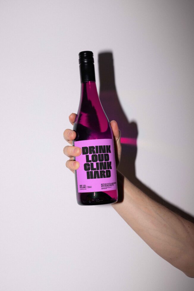 Hand holding a wine bottle with bold typographic label Drink loud clink hard vibrant design for mockup graphic template branding designers asset