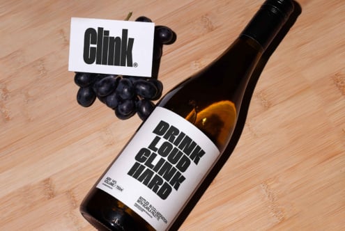 Wine bottle on wooden surface with bold label text Drink Loud Clink Hard, accompanied by grapes and card. Ideal for designers seeking typography inspiration.