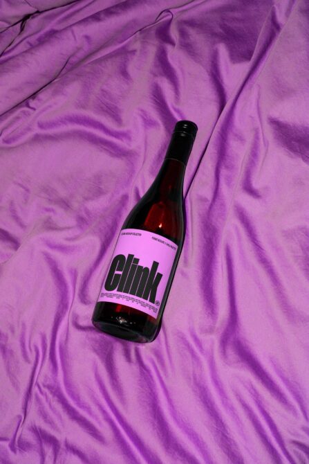 Dark bottle with pink Clink label on purple crumpled fabric, flat lay setup