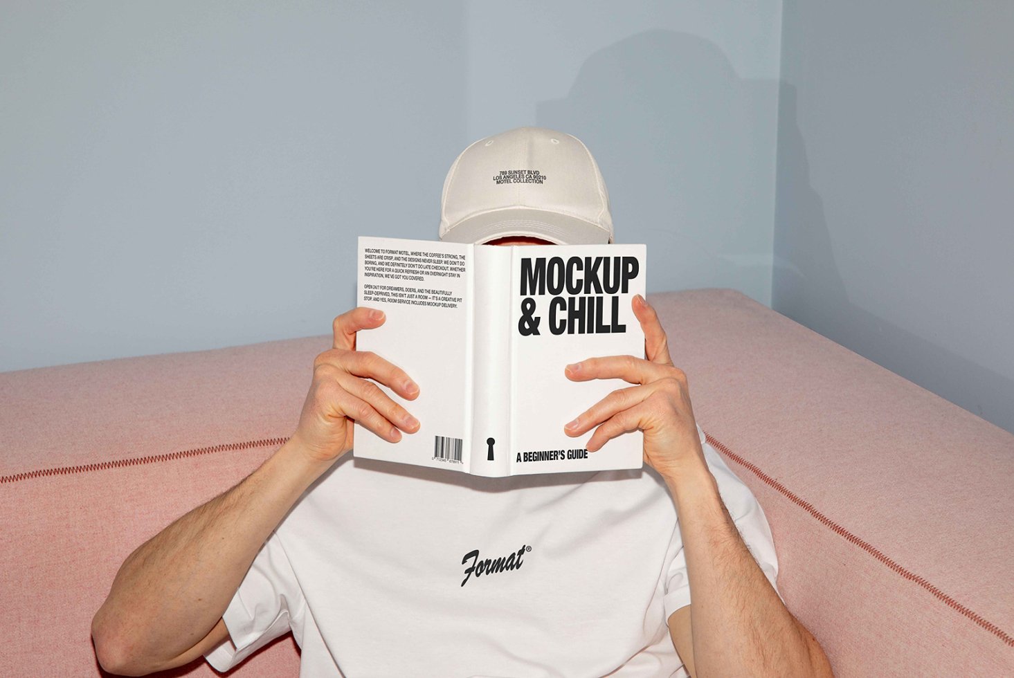 Man reading Mockup and Chill book, wearing white shirt and cap, seated on pink sofa. Perfect for design templates with modern casual style.