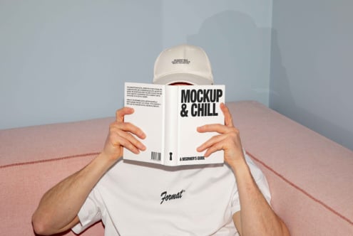 Man reading Mockup and Chill book, wearing white shirt and cap, seated on pink sofa. Perfect for design templates with modern casual style.