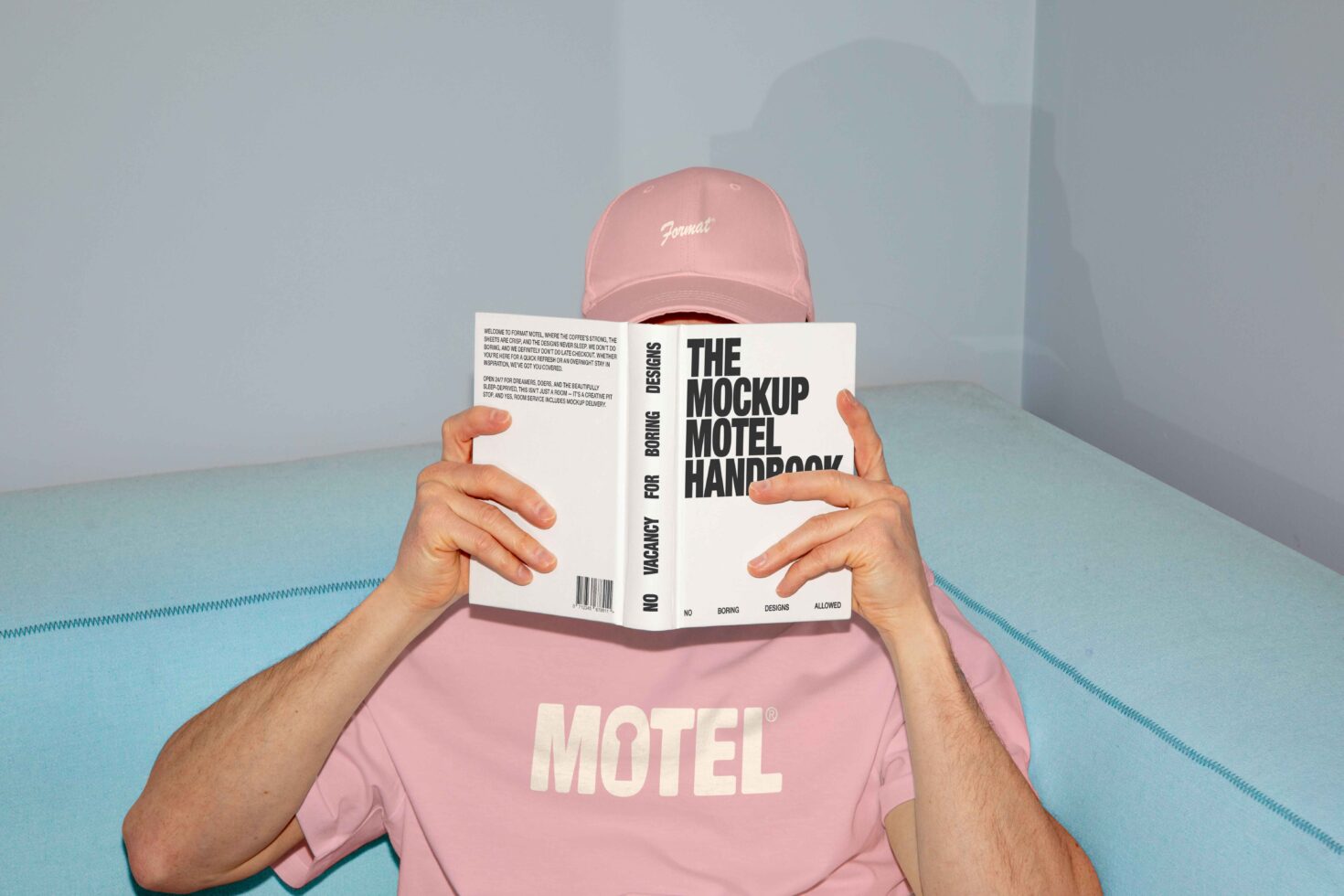 Designer in pink shirt and cap reading The Mockup Motel Handbook on a blue sofa. Perfect for showcasing design mockups and creative templates.
