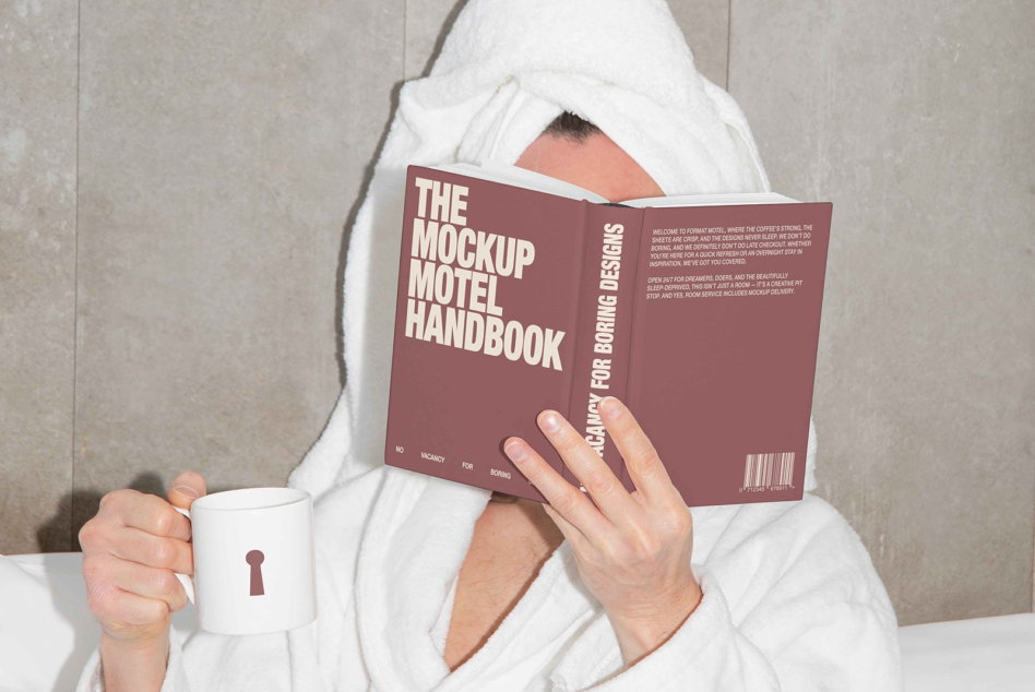 Person in white robe holding burgundy Mockup Motel Handbook with white mug
