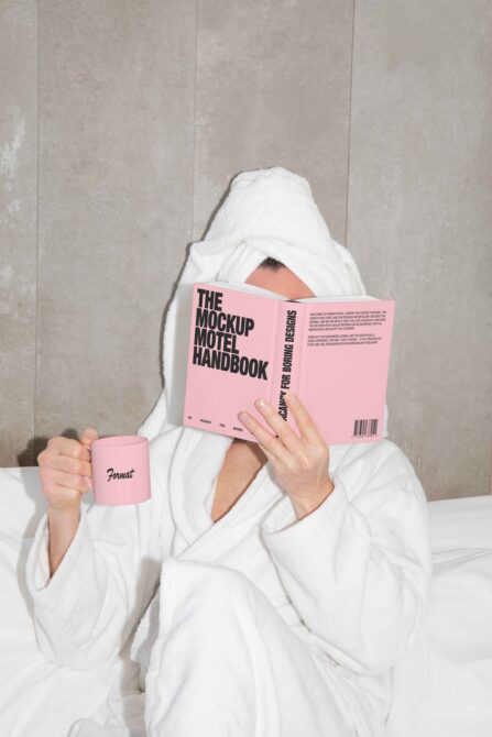 Person in white robe holding pink Mockup Motel Handbook with pink mug