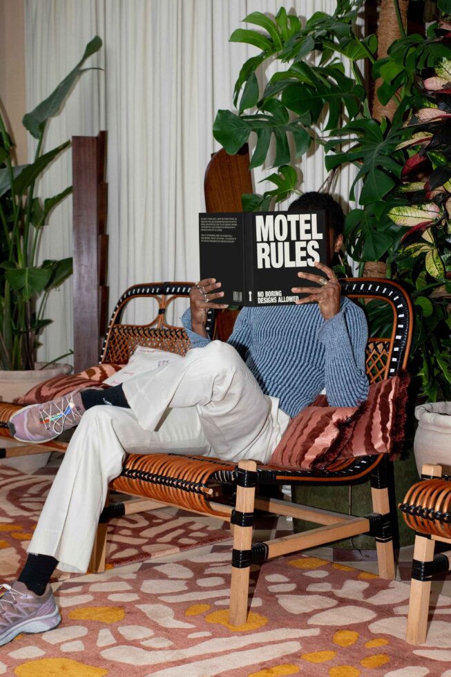 Person on patterned bench reads Motel Rules book, surrounded by lush plants; interior design inspiration, creative space, cozy atmosphere for designers.