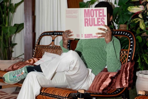 Person in green sweater reading book titled Motel Rules on a cozy wicker chair surrounded by plants. Perfect for graphic design inspiration mockup.