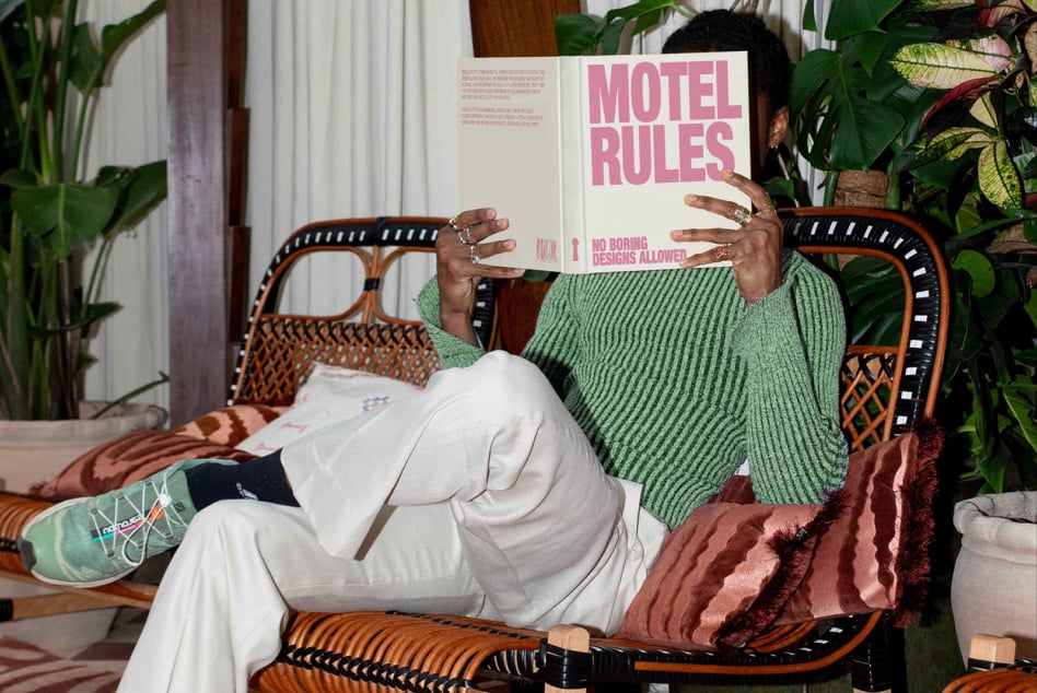 Person in green sweater sitting in woven chair holding pink Motel Rules magazine