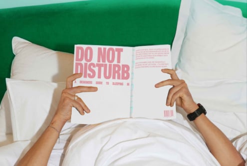 Person in bed reading a book titled Do Not Disturb A Designers Guide to Sleeping in. Green headboard pillows bed setting. Ideal for mockups or templates.