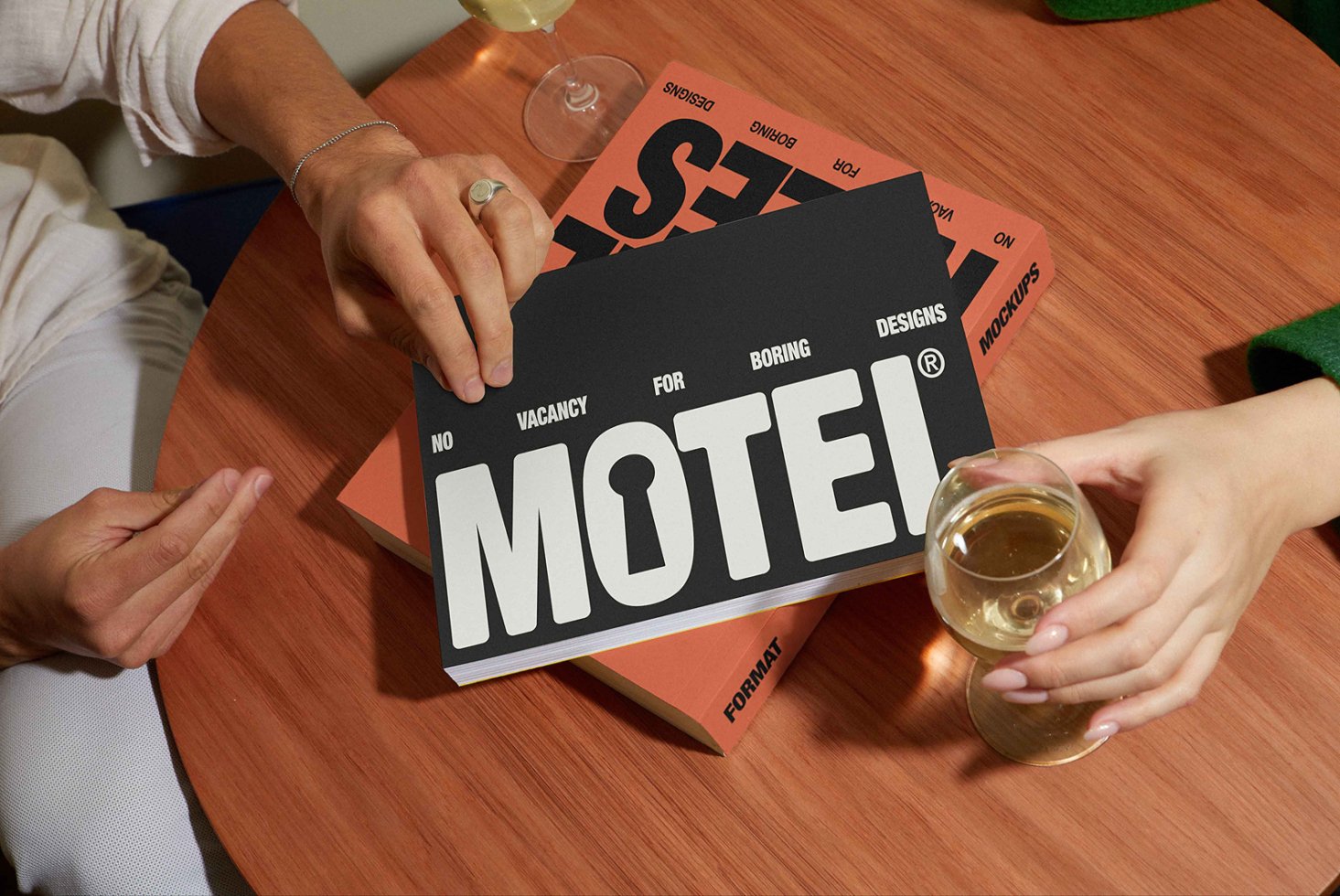 Designer holding black and orange mockup booklet with bold text No Vacancy For Boring Designs Motel next to wine glass on wooden table marketing templates