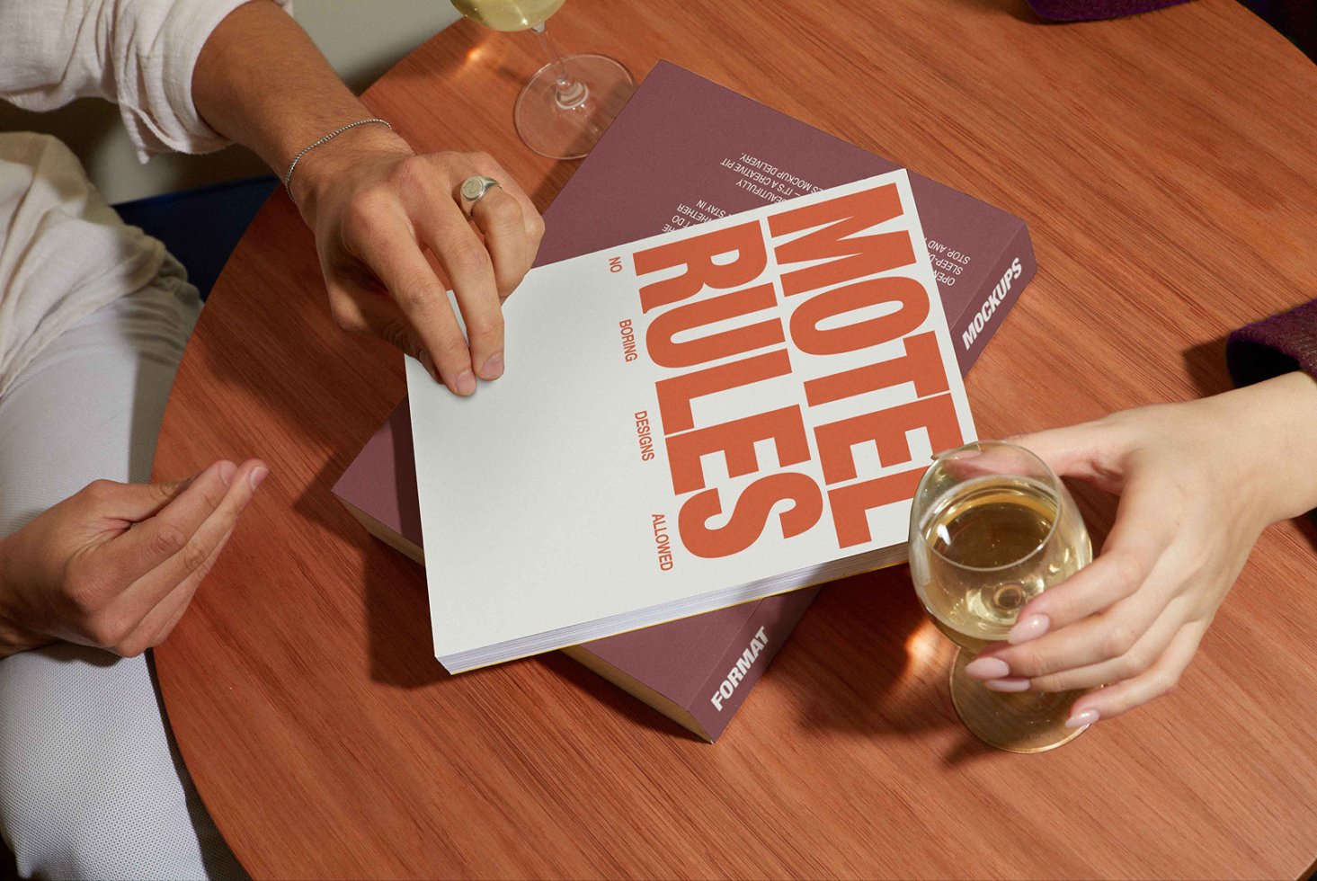 Two people interact with a graphic design mockup book on a wooden table with wine glasses. Keywords: mockup, graphic design, template, book, table