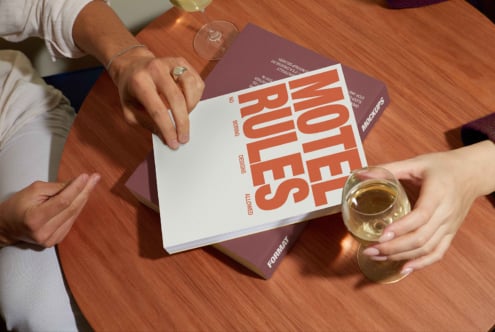 Two people interact with a graphic design mockup book on a wooden table with wine glasses. Keywords: mockup, graphic design, template, book, table