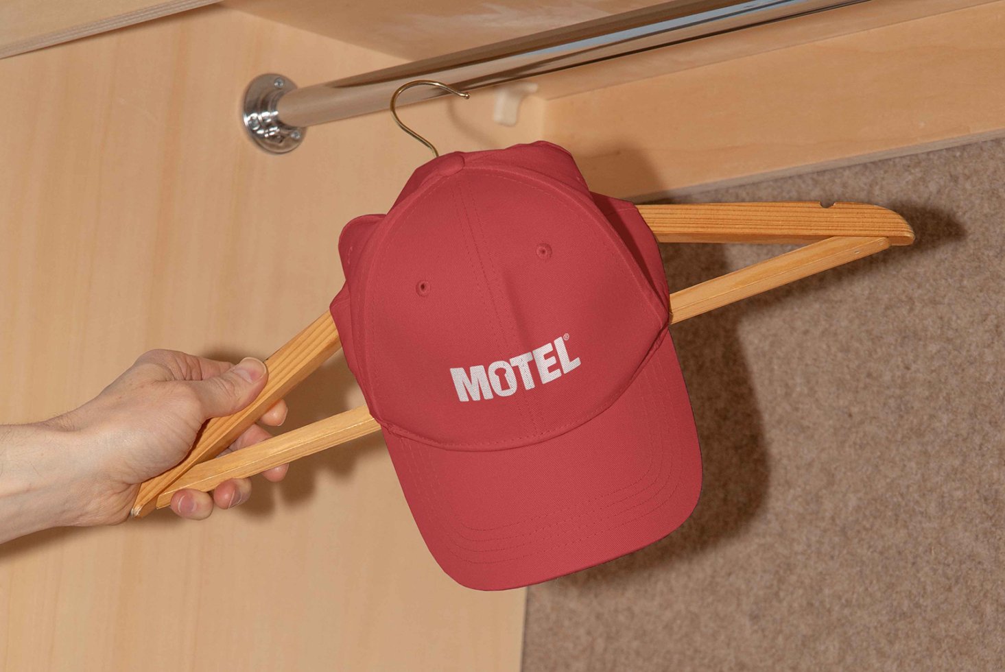 Red cap with white Motel logo hanging on wooden hanger in a closet setting. Ideal for apparel mockups for graphic designers and branding projects.