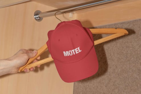 Red cap with white Motel logo hanging on wooden hanger in a closet setting. Ideal for apparel mockups for graphic designers and branding projects.