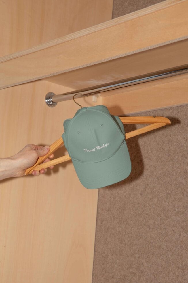 Hand holding a wooden hanger with a cap, customizable hat mockup, apparel design asset, wardrobe setting, tailored for designers, closet background.