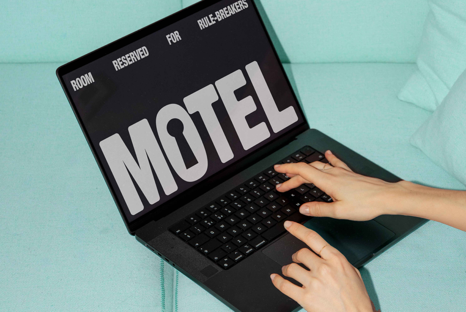 Laptop screen with cream motel sign on dark background, hands typing on mint couch