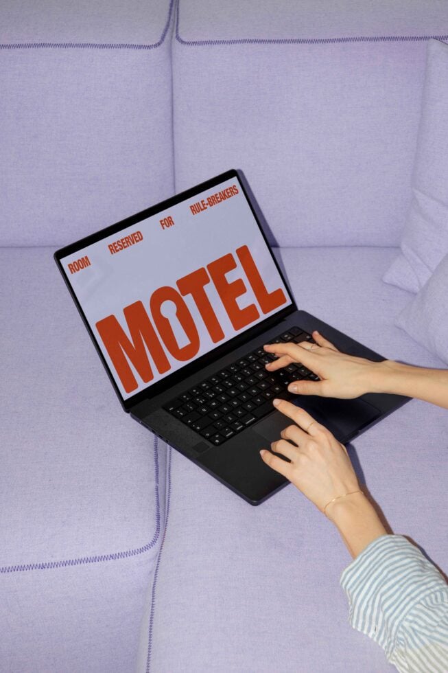 Laptop on purple sofa displaying Motel graphic design. Hands typing on keyboard. Keywords: graphic design, mockup, laptop, typography, designers asset.