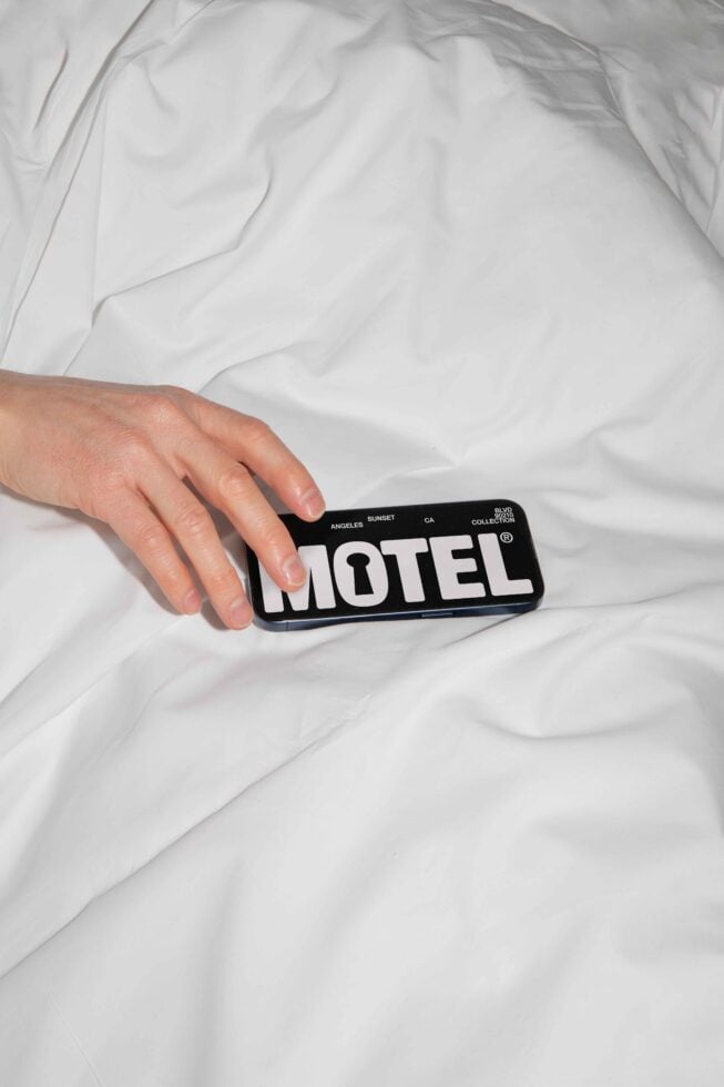 Hand holding a phone with Motel text on screen on white bed sheets background suitable for mockup design, graphic assets, digital templates for designers.