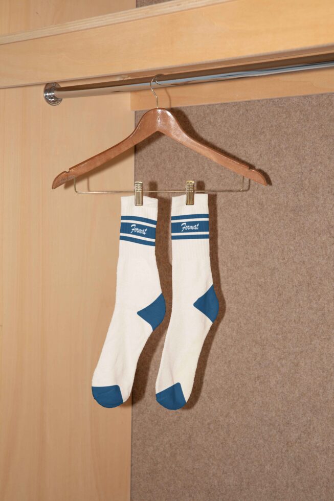 Pair of white socks with blue accents on a wooden hanger in a closet setting. Perfect for apparel mockup design templates for designers.