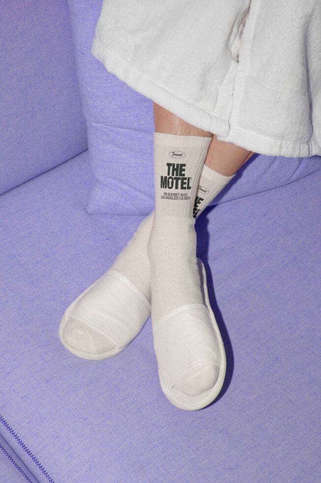 Person in cozy white robe and socks with The Motel branding relaxes on a soft lavender couch. Ideal for mockup, template, and graphic design use.