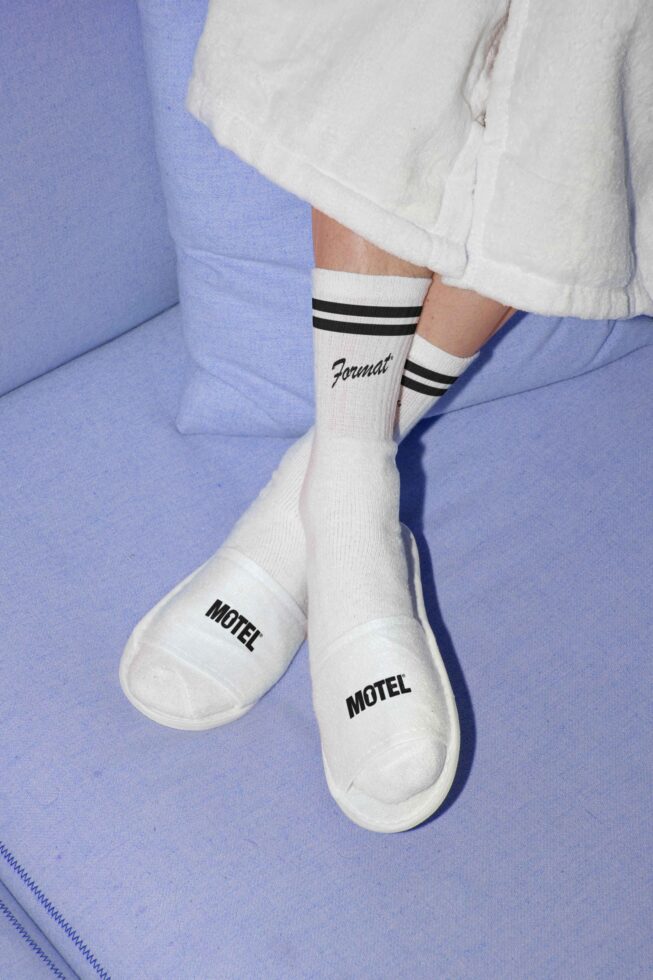 Casual mockup featuring person wearing white socks and matching branded slides on a light blue couch. Perfect for fashion graphics or branding templates.
