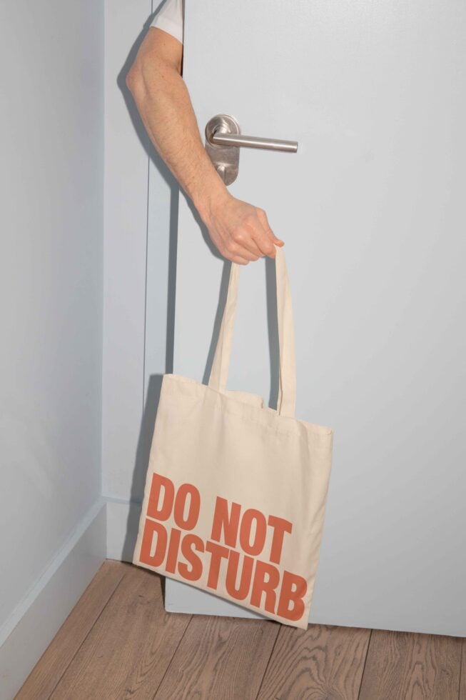Hand holding a tote bag with Do Not Disturb text, ideal for mockups. Door background, minimalist style, suitable for designers seeking unique graphics.