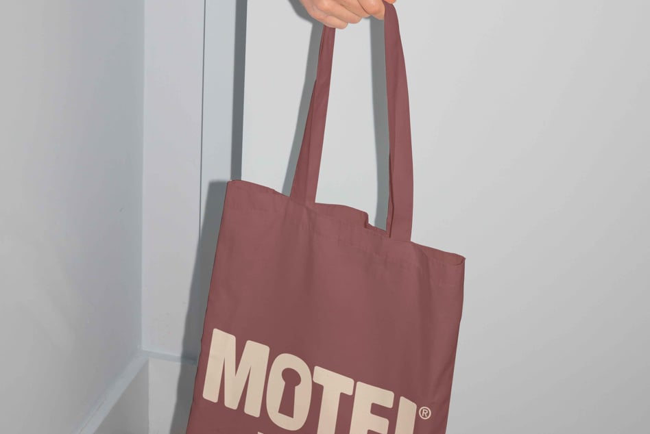 Person carrying burgundy tote with cream Motel lettering in minimal space