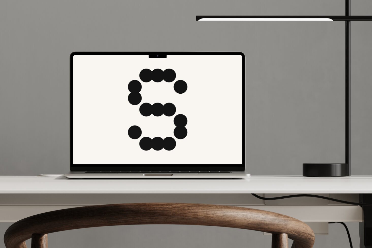 Laptop displaying bold circular graphic letter S on minimal white desk with wooden chair and modern black lamp. Ideal for office mockup designers.