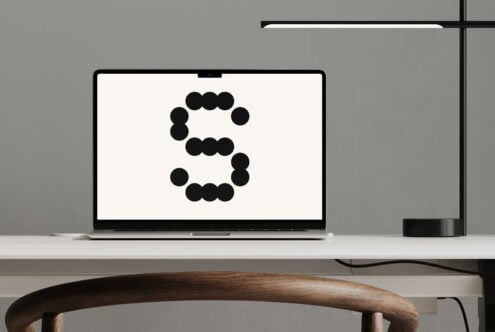 Laptop displaying bold circular graphic letter S on minimal white desk with wooden chair and modern black lamp. Ideal for office mockup designers.