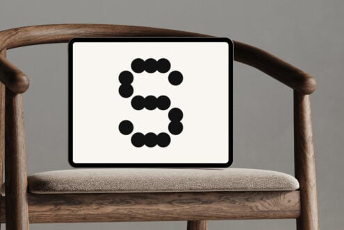 Tablet on wooden chair displaying letter S formed with black dots on a white screen. Minimalistic design ideal for font or mockup templates for designers.