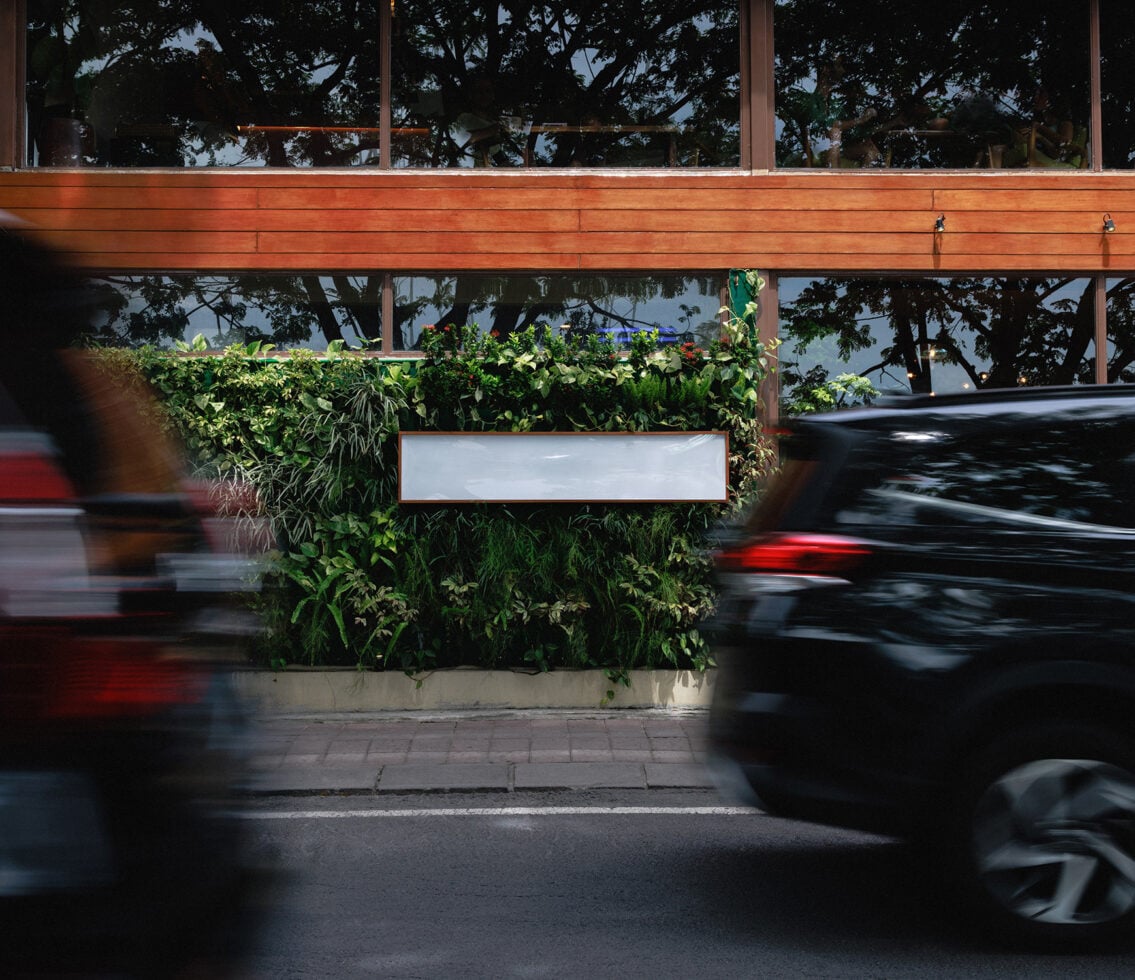 Blank storefront sign on lush green planter wall with blurred cars passing by