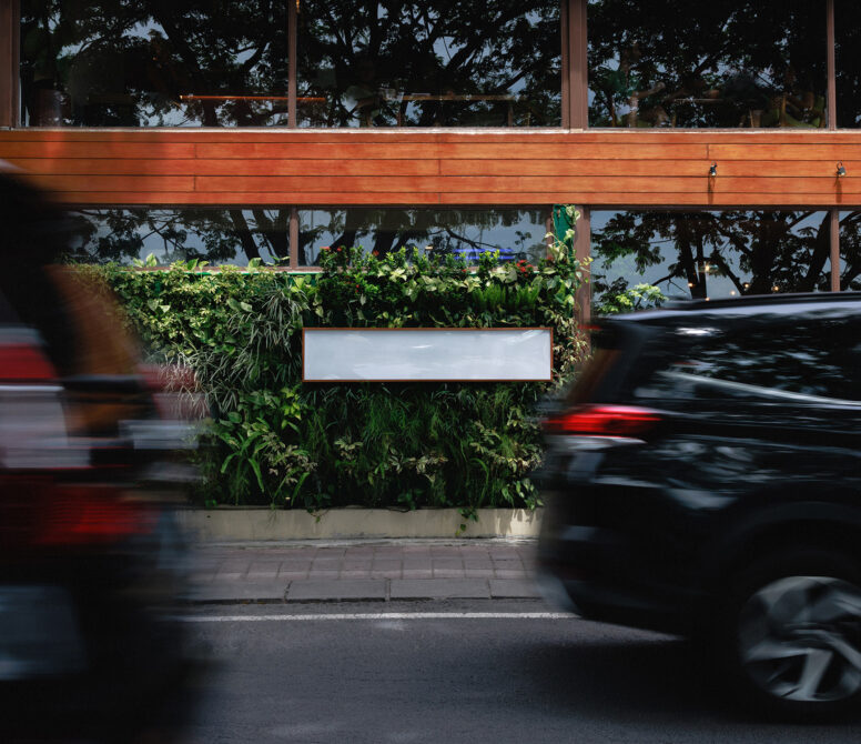 Blank storefront sign on lush green planter wall with blurred cars passing by