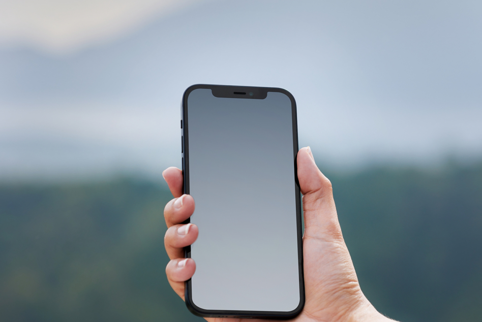 Hand holding iPhone with blank gray screen against blurred outdoor landscape background