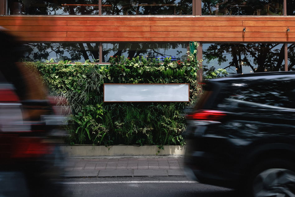 Empty horizontal sign mounted on verdant plant wall with moving vehicles on street