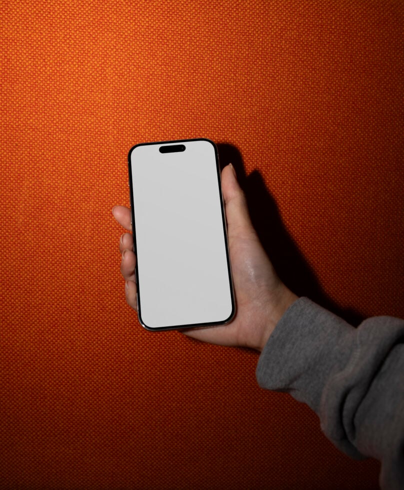 Mockup of a smartphone held by a hand in front of an orange background. Blank screen for app design display. Ideal for digital assets and templates.