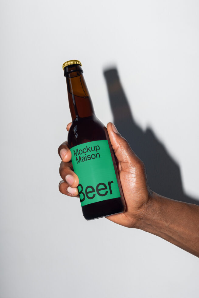 Hand holding a beer bottle with a green label reading Mockup Maison Beer. Perfect for packaging mockups. Shadow on white background. Ideal for designers.