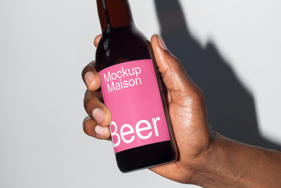 Hand holding dark beer bottle with bright pink label showing Mockup Maison branding