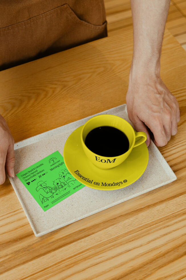Coffee cup and saucer set on table next to a small green card. Ideal for mockup designs showcasing branding, kitchen themes, or coffee-related graphics.
