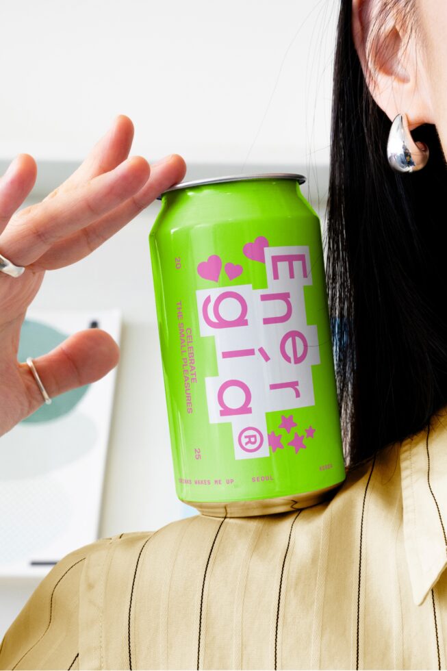 Close-up of a vibrant green energy drink can with pink typography, held near a person's ear, perfect for branding mockups and designer assets.