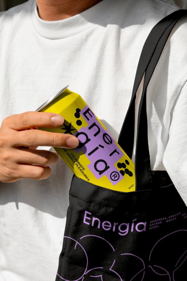 Person holding a yellow energy drink can with modern design near a black tote bag featuring bold text. Ideal for packaging mockups and branding insights.