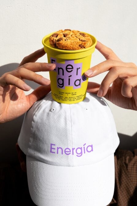 Person wearing white cap with purple Energia text holds yellow cup with a cookie. Ideal for branding mockup, product display, packaging design template.