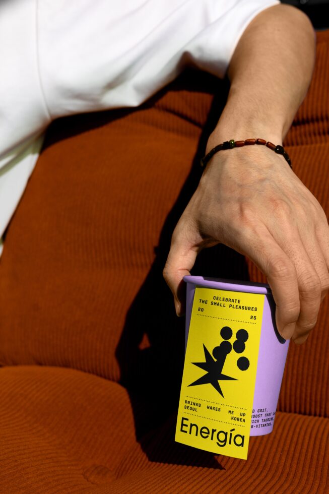 Hand holding a purple cup with yellow graphic label on orange fabric; ideal for product mockups, packaging design, branding, creative templates.