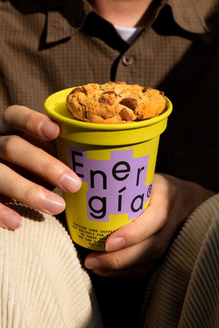 Close-up of a hand holding a vibrant yellow coffee cup labeled Energia topped with a cookie. Perfect for mockup design or branding template showcase.