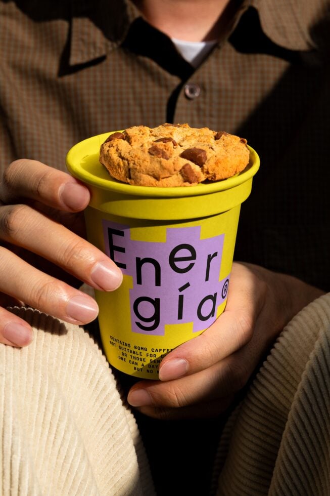 Close-up of a hand holding a vibrant yellow coffee cup labeled Energia topped with a cookie. Perfect for mockup design or branding template showcase.