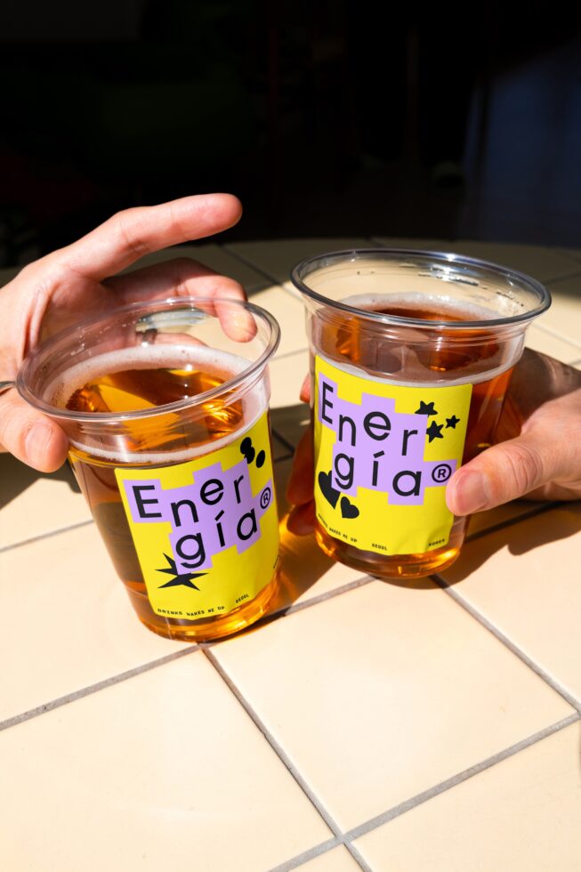 Hands hold plastic cups labeled Energia with vibrant yellow and purple graphics containing iced tea on a tiled surface. Perfect for branding mockup design.