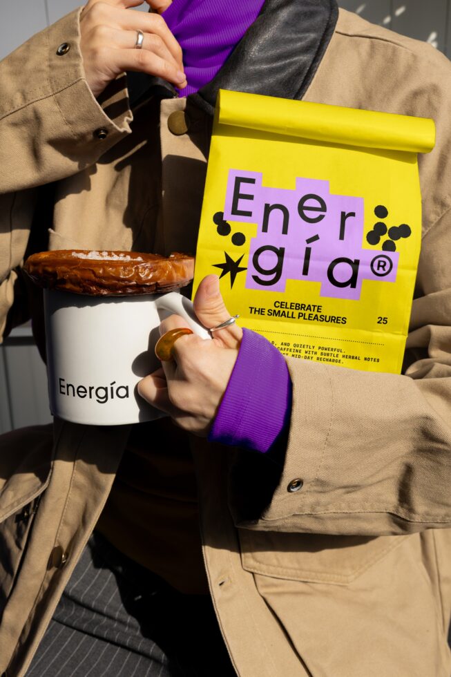 Person holding a mug and a yellow package of coffee labeled Energia wearing a tan coat and purple sleeve. Ideal for designers seeking branding inspiration.