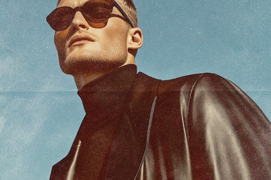 Man in brown leather jacket wearing oversized sunglasses against blue sky