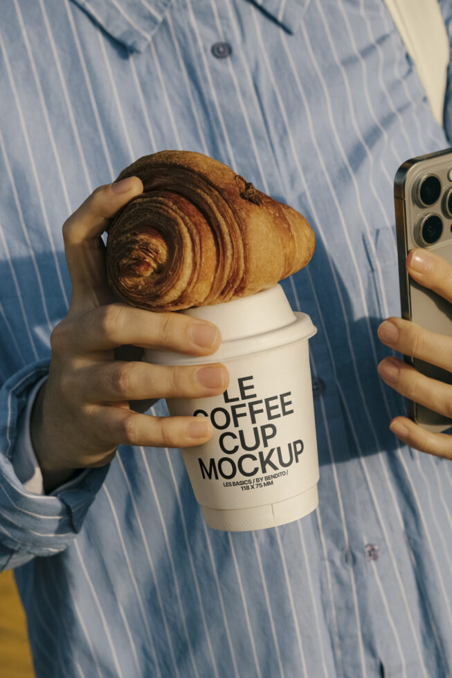Person holds a coffee cup mockup and croissant in one hand and a smartphone in the other wearing a blue striped shirt design asset for designers