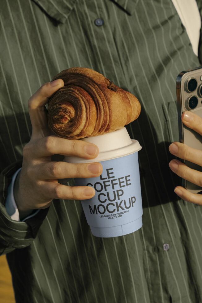 Person holding a smartphone and blue coffee cup with croissant. Cup reads Le Coffee Cup Mockup. Ideal for designers seeking realistic templates mockups.