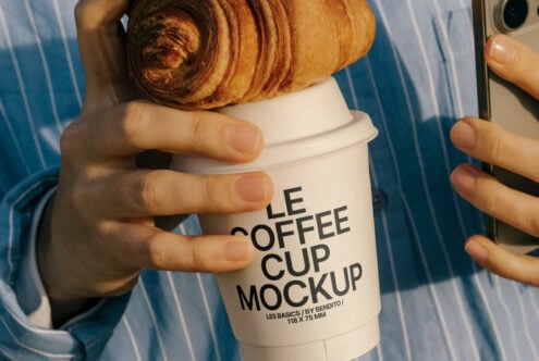 Hand holds croissant and coffee cup with Le Coffee Cup Mockup text, wearing blue striped shirt, ideal for design mockups, cafe branding, packaging.